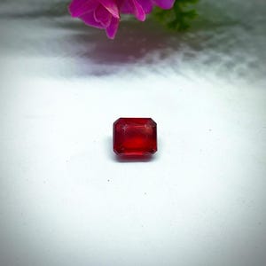 Può includere: Una gemma rossa vibrante, a taglio quadrato, con una superficie sfaccettata. La gemma è posta su uno sfondo bianco, con un accenno di fiori rosa nell'angolo in alto a sinistra. Il colore della gemma è un rosso intenso e ricco.