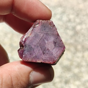 Peut inclure: Une pierre précieuse rubis brute et non taillée avec une teinte rouge violacé profond. La pierre présente une forme hexagonale avec des structures cristallines internes visibles, des facettes naturelles et un aspect semi-translucide.
