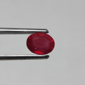 Peut inclure: Un rubis de forme ovale, d'un rouge profond, maintenu par une pince argentée. La pierre précieuse présente des nuances de couleur subtiles. Le rubis est mis en valeur sur un fond clair et uni.