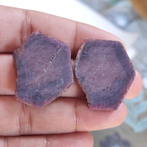 May include: Two raw, hexagonal-shaped ruby gemstones with a deep purplish-red hue. The stones feature a matte, textured surface with visible natural inclusions and geometric patterns across their flat, polished faces, showcasing a dark colour.