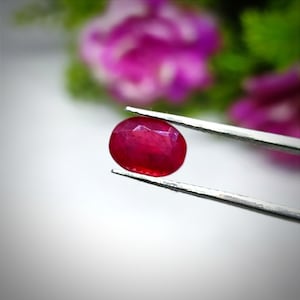 Könnte beinhalten: Ein leuchtend roter ovaler Edelstein, der mit einer Pinzette gehalten wird. Der Edelstein hat eine glatte, polierte Oberfläche und eine tiefe, satte rote Farbe.