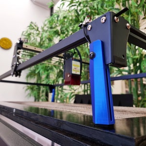 May include: A black and blue laser cutter with a black metal frame and blue metal legs. The laser cutter is sitting on a black surface.
