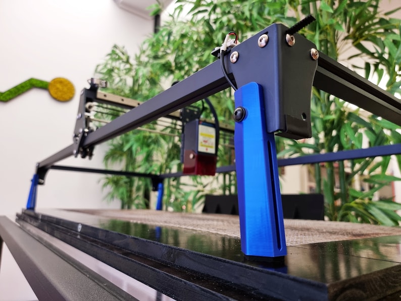 May include: A black and blue laser cutter with a black metal frame and blue metal legs. The laser cutter is sitting on a black surface.