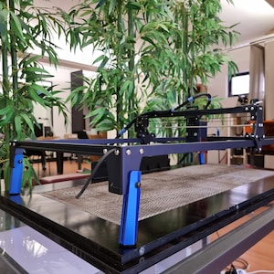 May include: A black 3D printer with a blue frame and blue supports. The printer is on a glass table with a black surface.