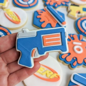 May include: A blue and orange Nerf gun cookie decorated with white icing. The cookie is held in a hand.