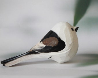 May include: A white wooden bird figurine with black and brown painted details. The bird is perched on a white surface with a blurred green background.