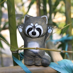 Puede incluir: Una figurita de mapache de madera con ojos negros y cara blanca, de pie con los brazos levantados. La figurita está pintada de gris y blanco y se coloca sobre un fondo de bambú verde.