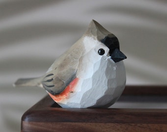 May include: A hand-carved wooden bird figurine with a gray and white body, black eyes, and a black beak. The bird has a small orange patch on its chest and is perched on a dark brown wooden surface.