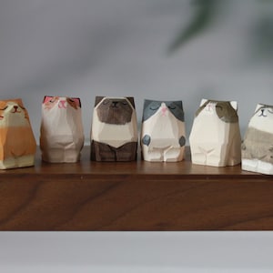 Puede incluir: Seis figuritas de madera de gato en varios colores y poses. Los gatos están todos sentados y tienen diferentes expresiones faciales. Las figuritas están dispuestas en una fila en un estante de madera.