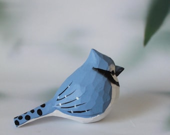 May include: A hand-carved wooden blue jay figurine. The bird features a light blue body with black and white accents, including a black beak and wing markings. The tail has black spots.