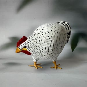 May include: A white and black chicken figurine with a red comb and yellow beak. The chicken is standing on a white surface.