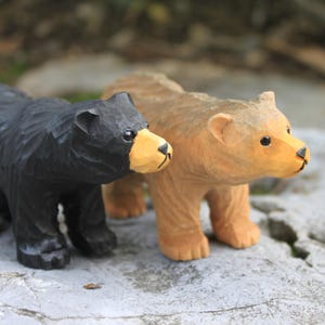 May include: Two wooden bear figurines, one black and one brown, standing side by side on a gray surface.
