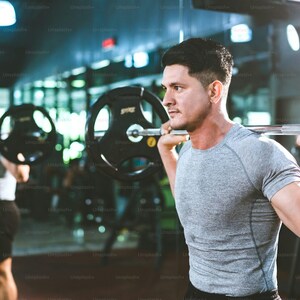 May include: A person wearing a grey short-sleeved athletic shirt performing a barbell back squat exercise in a gym. The barbell features black weight plates on each side, and the person is positioned with the bar resting across their shoulders.