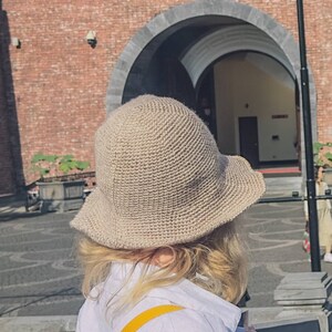 May include: A person wearing a beige crocheted bucket hat with a wide brim. The hat is made of a textured material and has a simple design.