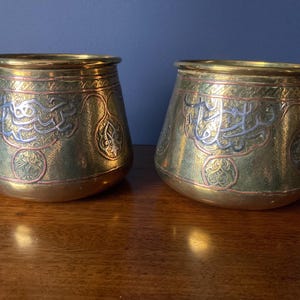 May include: Two antique brass vessels with a gold finish, featuring intricate silver and copper-colored etched designs. The vessels have a rounded base and a wider top, with Arabic calligraphy. The vessels are displayed on a wooden surface.