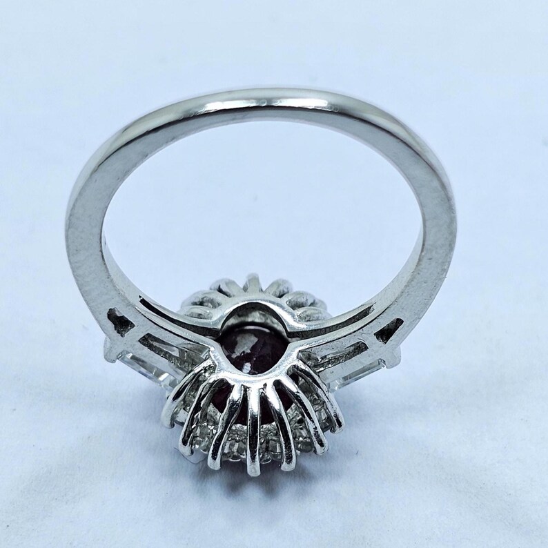 Puede incluir: Un anillo plateado con una piedra central redonda de color rojo oscuro, rodeada de piedras m&aacute;s peque&ntilde;as y transparentes. La banda del anillo es pulida, con piedras transparentes a cada lado del grupo central. El anillo est&aacute; sobre un fondo claro.
