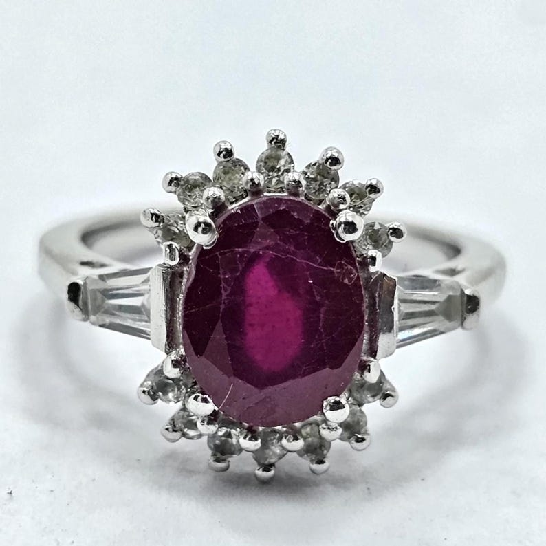 Puede incluir: Un anillo de plata con una gran gema de rub&iacute; de corte ovalado y piedras de acento m&aacute;s peque&ntilde;as. El rub&iacute; es de color rojo intenso, y las piedras de acento son claras. El anillo tiene un dise&ntilde;o cl&aacute;sico y elegante.