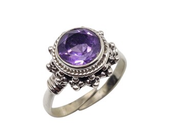 May include: A silver ring featuring a large, round, faceted purple gemstone. The gemstone is set in a decorative bezel with a rope-like border and small, beaded accents. The ring's band is adjustable.