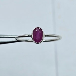 Op de afbeelding: Een zilveren ring met een ovaalvormige, dieprode robijn. De robijn is gezet in een eenvoudige band. De ring wordt getoond tegen een witte achtergrond.