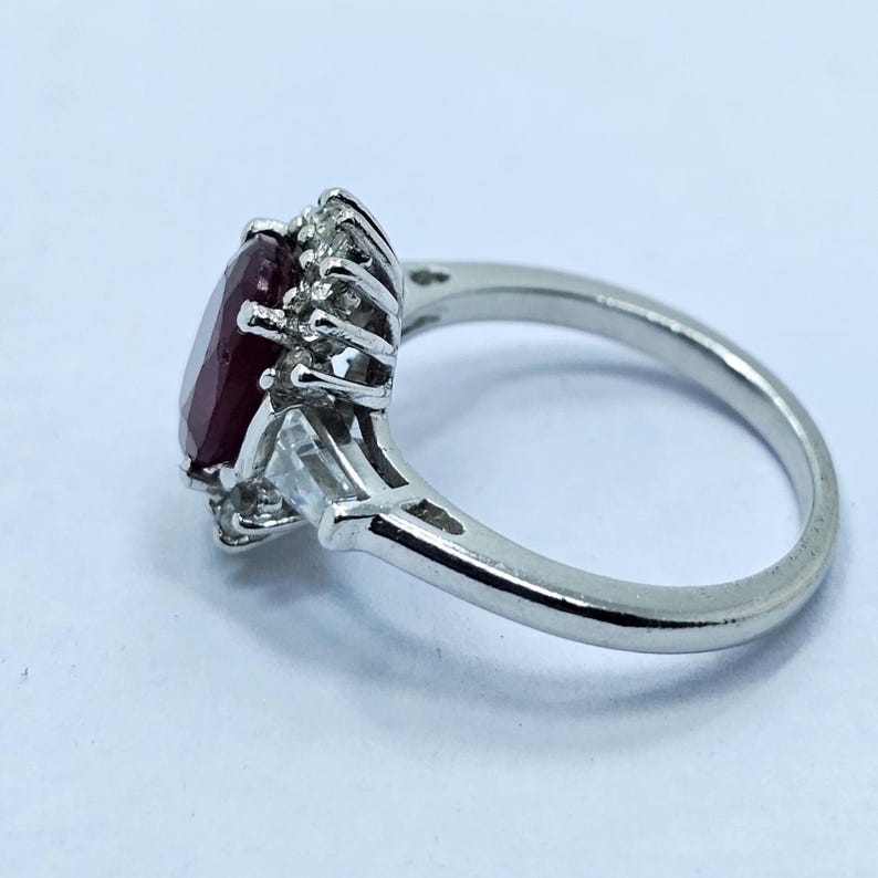 Puede incluir: Un anillo de plata con una gran gema roja oscura de talla ovalada, rodeada de piedras claras m&aacute;s peque&ntilde;as. El anillo tiene un dise&ntilde;o cl&aacute;sico con una banda pulida y un engaste de garras para la piedra central. El anillo est&aacute; sobre un fondo blanco.