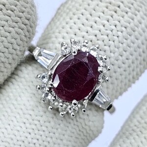 Puede incluir: Un anillo de plata con un gran rub&iacute; ovalado, rodeado de piedras claras m&aacute;s peque&ntilde;as. Dos piedras de talla baguette flanquean el rub&iacute;. El anillo est&aacute; sobre un fondo blanco.