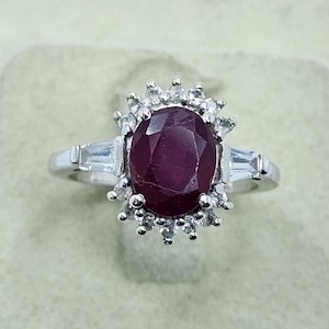 Puede incluir: Anillo de plata con un gran rub&iacute; de corte ovalado. El rub&iacute; est&aacute; rodeado de piedras preciosas claras m&aacute;s peque&ntilde;as. Dos piedras de talla baguette flanquean el rub&iacute;. El anillo tiene un dise&ntilde;o cl&aacute;sico y elegante.
