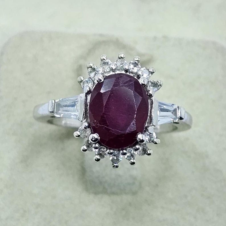 Puede incluir: Anillo de plata con un gran rub&iacute; de corte ovalado. El rub&iacute; est&aacute; rodeado de piedras preciosas claras m&aacute;s peque&ntilde;as. Dos piedras de talla baguette flanquean el rub&iacute;. El anillo tiene un dise&ntilde;o cl&aacute;sico y elegante.