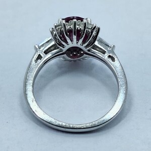 Puede incluir: Un anillo plateado con una piedra preciosa grande, redonda y de color rojo oscuro, rodeada de piedras claras m&aacute;s peque&ntilde;as. Dos piedras claras rectangulares est&aacute;n engastadas a cada lado de la piedra central. El anillo est&aacute; sobre un fondo claro.