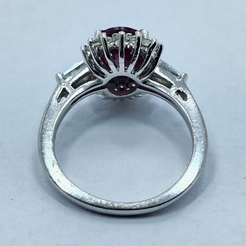 Puede incluir: Un anillo plateado con una piedra preciosa grande, redonda y de color rojo oscuro, rodeada de piedras claras m&aacute;s peque&ntilde;as. Dos piedras claras rectangulares est&aacute;n engastadas a cada lado de la piedra central. El anillo est&aacute; sobre un fondo claro.