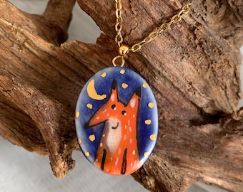 May include: A gold-colored chain necklace with a pendant featuring a cartoon fox illustration. The oval pendant has a blue background with a crescent moon and stars. The fox is orange and white.