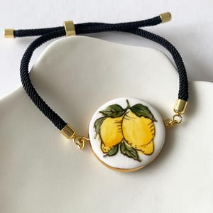 Könnte beinhalten: Schwarzes Kordelarmband mit einem runden, weißen Keramikanhänger mit einem handgemalten Design von zwei gelben Zitronen mit grünen Blättern. Der Anhänger ist goldfarben umrandet, und das Armband hat goldfarbene Metallakzente.