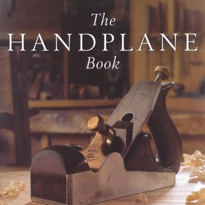 Könnte beinhalten: Das Buchcover von The Handplane Book zeigt einen Hobel mit Holzgriff und Metallklinge. Der Titel ist in weißer Schrift. Der Hobel liegt auf einer Holzoberfläche, daneben Holzspäne. Auf dem Hobel steht "Norris A6 London".