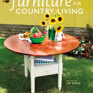 May include: A round wooden table with a dark brown top and white base, set on a green lawn. The table is laden with a green bowl of fruit, vegetables, and a vase of sunflowers. The book cover reads "Building Furniture for Country Living."
