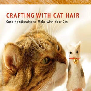 Può includere: Un gatto tigrato e bianco guarda un burattino a dito a forma di gatto in feltro. Il burattino ha un collare rosso con una campana dorata e macchie nere. Il testo recita "Crafting with Cat Hair" e "Cute Handicrafts to Make with Your Cat".