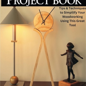 Pode incluir: Um livro intitulado "The Biscuit Joiner Project Book" com um relógio de madeira em um suporte e uma figura de bronze de uma menina em uma base de madeira. A capa do livro apresenta o título em letras grandes e em negrito e um subtítulo que diz "Dicas e técnicas para simplificar seu trabalho de madeira usando esta ótima ferramenta".