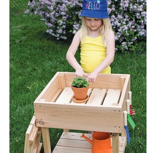 May include: A wooden potting bench with a small terracotta pot containing a green plant. An orange watering can sits below. A child wearing a blue hat with the word "HELLO" is tending to the plant. The bench is on a grassy lawn.