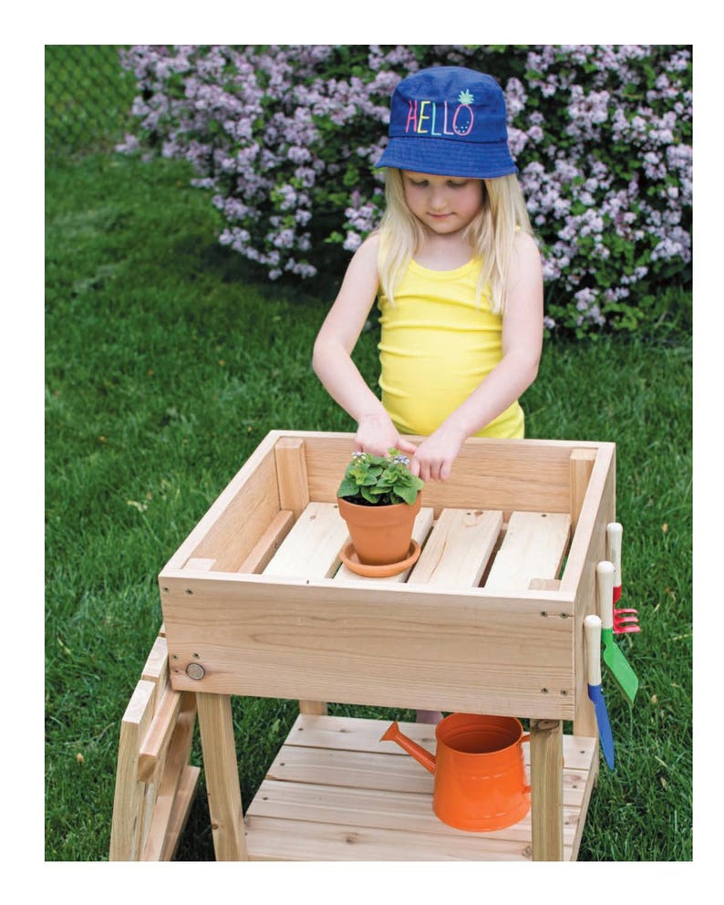 May include: A wooden potting bench with a small terracotta pot containing a green plant. An orange watering can sits below. A child wearing a blue hat with the word "HELLO" is tending to the plant. The bench is on a grassy lawn.