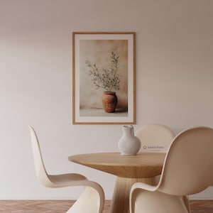 May include: A framed wall art print featuring a terracotta pot with green olive branches against a neutral, textured background. The print is displayed in a light wood frame, hanging on a plain wall above a round wooden table with chairs.