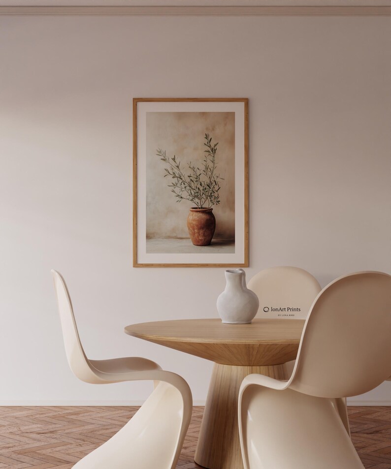 May include: A framed wall art print featuring a terracotta pot with green olive branches against a neutral, textured background. The print is displayed in a light wood frame, hanging on a plain wall above a round wooden table with chairs.