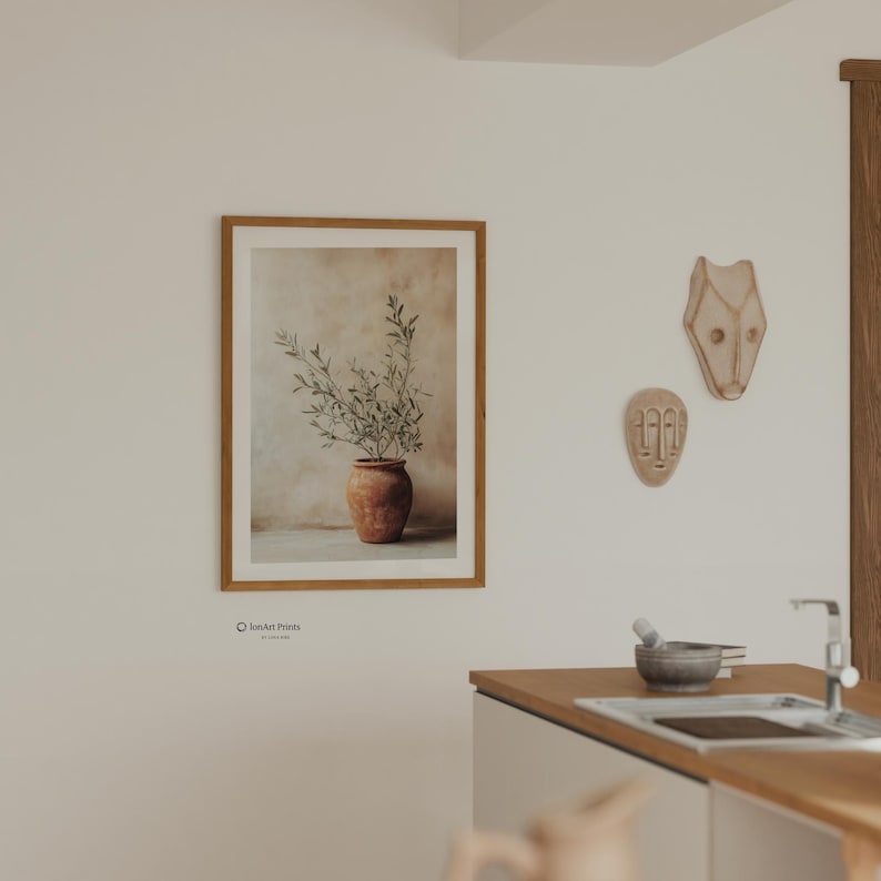 May include: A framed wall art print featuring a terracotta-colored pot containing a green olive branch against a beige background. The print is displayed on a wall next to two decorative masks and a kitchen counter with a sink and faucet.
