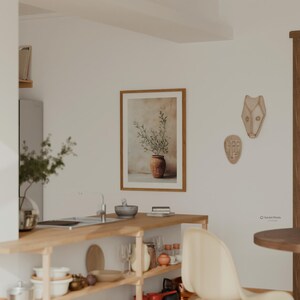 May include: A framed wall art print featuring a terracotta pot with olive branches, displayed on a white wall next to two abstract face masks. The kitchen scene includes a wooden counter with various kitchenware, including bowls and glasses.