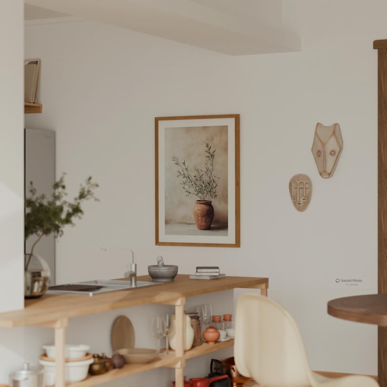 May include: A framed wall art print featuring a terracotta pot with olive branches, displayed on a white wall next to two abstract face masks. The kitchen scene includes a wooden counter with various kitchenware, including bowls and glasses.