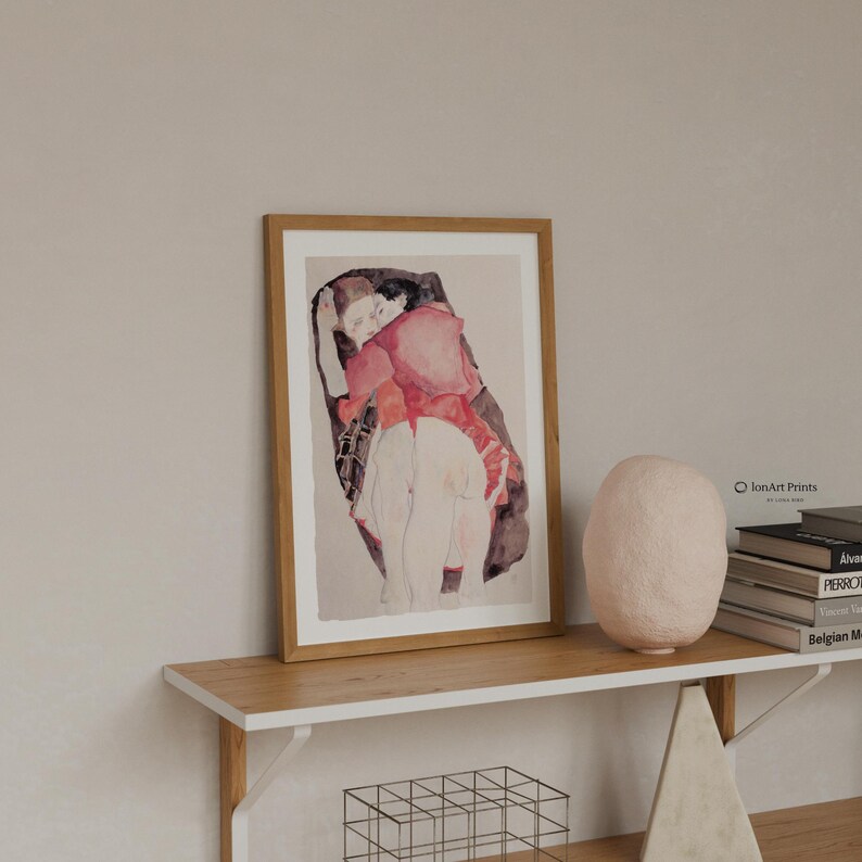 Puede incluir: Una l&aacute;mina art&iacute;stica enmarcada con una ilustraci&oacute;n en acuarela de dos figuras abrazadas, en tonos rojos, negros y beige. La obra est&aacute; expuesta en un marco de madera clara, sobre un estante de madera junto a una escultura de color rosa p&aacute;lido.