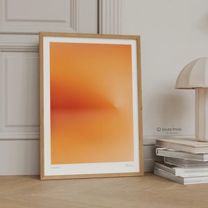 May include: A framed art print featuring an abstract gradient of orange and red hues, evoking a sunset. The artwork is encased in a light brown wooden frame. The print is labeled "Sunset" at the bottom. The artist's logo is visible in the lower right corner.