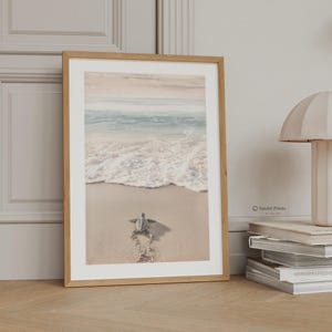 May include: A framed wall art print featuring a small sea turtle crawling across light beige sand toward the ocean waves. The scene is captured in soft, muted tones with the text IonArt Prints visible on the lower right side of the print.