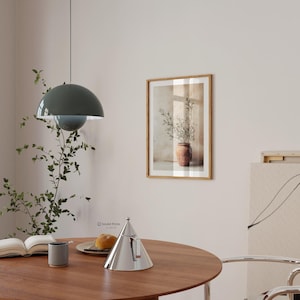 May include: A framed wall art print featuring a terracotta pot with olive branches, displayed above a round wooden table with a silver conical teapot, a gray mug, an open book, and a plate with bread, under a dark green dome pendant light.