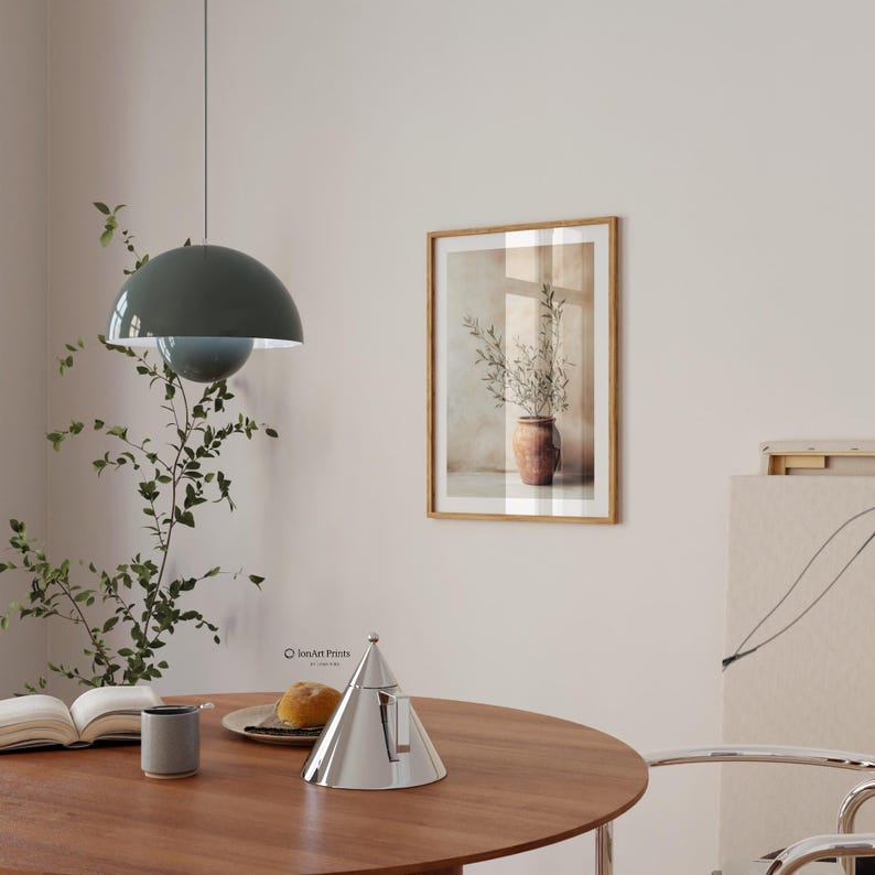 May include: A framed wall art print featuring a terracotta pot with olive branches, displayed above a round wooden table with a silver conical teapot, a gray mug, an open book, and a plate with bread, under a dark green dome pendant light.