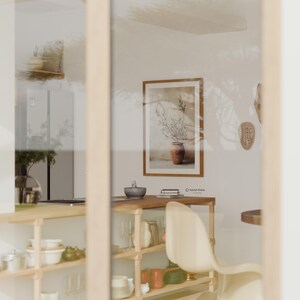 May include: A framed wall art print featuring a botanical illustration of an olive branch in a terracotta pot, displayed in a kitchen interior with wooden shelving, a white chair, and a small mortar and pestle on a wooden counter surface.