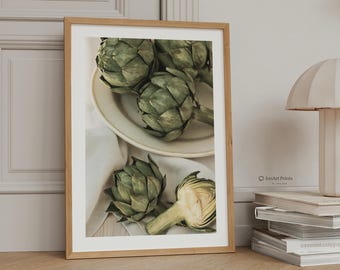 May include: A framed wall art print featuring a high-angle photograph of several green artichokes. Two whole artichokes rest on a cream-colored plate, while another whole artichoke and a halved artichoke sit on a white cloth below the plate.