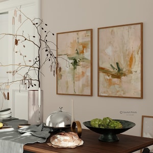 Op de afbeelding: Twee ingelijste abstracte kunstprints met neutrale tinten beige, groen en oranje. Het kunstwerk wordt tentoongesteld op een lichtbeige muur. Een tafel is gedekt met een decoratieve tak, een zilveren cloche en een kom met artisjokken.
